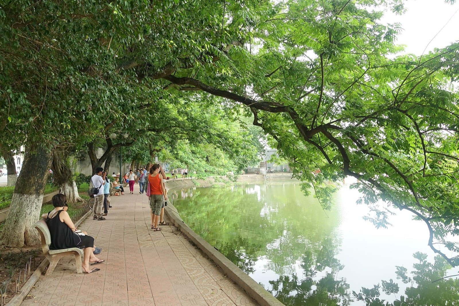 A tree-shaded lakeside path along Hoan Kiem Lake in central Hanoi, with walkers and stone benches